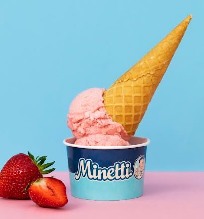 Vaaleanpunainen mansikkaj&auml;&auml;tel&ouml;pallo sinisess&auml; Minetti-j&auml;&auml;tel&ouml;kupissa, jonka p&auml;&auml;ll&auml; on yl&ouml;salaisin asetettu vohvelikeksi. Kupin vieress&auml; on kokonainen ja puoliksi leikattu mansikka vaaleanpunaisella ja sinisell&auml; taustalla.