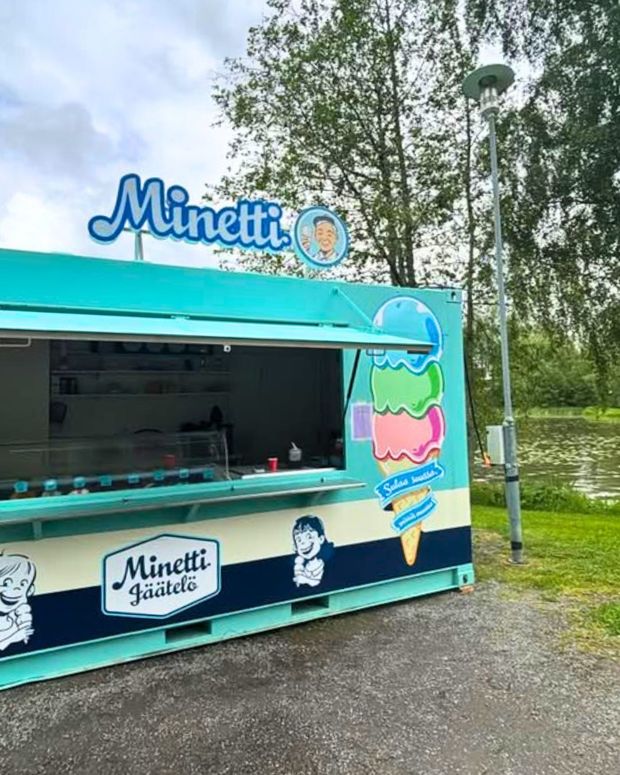 Minetti-jäätelökoju ulkona puiden keskellä. Koju on turkoosin ja valkoisen sävyinen, koristeltu piirretyillä lapsihahmoilla ja värikkäällä jäätelögrafiikalla. Taustalla näkyy pilvinen taivas ja hiukan järveä