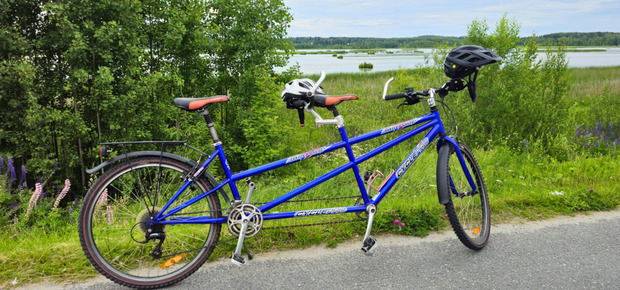 sininen tandempyörä pyörätien reunassa. Taustalla lehtipuita, ruohikkoa ja järvi
