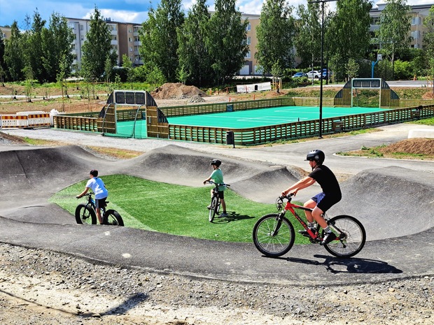 Three cyclists ride on the pumptrack at Halkola Sports Park in Lempäälä, with a multi-purpose sports field and surrounding park area in the background.