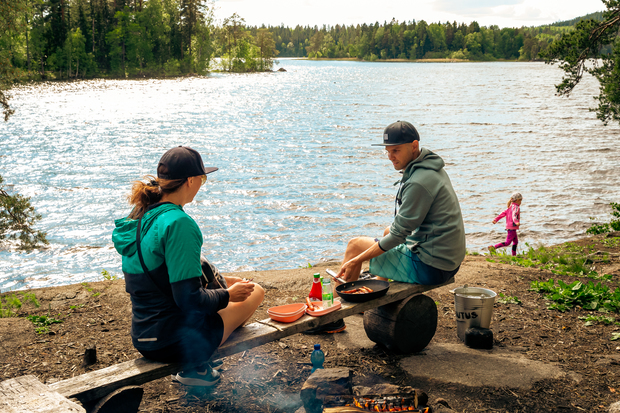 Two people sitting on a rock by a lakeside campfire, with a child walking in the background.