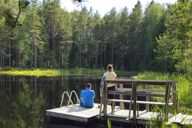 Two people on a small wooden pier by a calm forest lake with trees in the background.