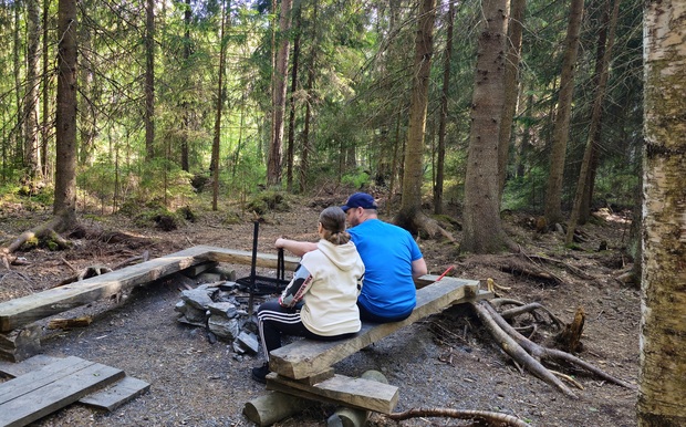 Kaksi henkilöä istuu metsässä Houkkammin nuotiopaikalla puisella penkillä tulipaikan äärellä. Heidän edessään on kivistä rakennettu nuotiopaikka ja ympärillä tiheää kuusimetsää, johon valo siivilöityy puiden välistä. Tunnelma on rauhallinen ja luonnonläheinen.
