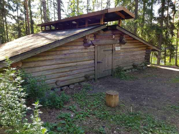 Hirsinen laavukota metsässä, edessä pieni puinen istuin ja sorapohjainen piha-alue. Katossa on tuuletusrakenne ja oven yläpuolella kyltti.