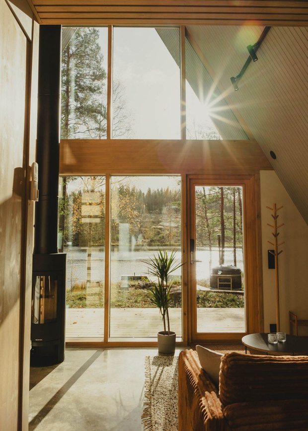 The bright interior of an A-frame cabin with large windows overlooking a lake; sunlight streams in, with a modern stove on the left and a cozy sofa on the right.