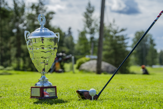 Hopeinen golfpalkintopokaali ja tiille asetettu golfpallo mailoineen nurmikolla, taustalla puustoa ja golfkentän maisemaa.
