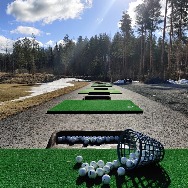 Golfpalloja kaatuneessa korissa ja lyöntialustoja jonossa ulkorangella, taustalla metsäinen maisema ja kevätaurinko.